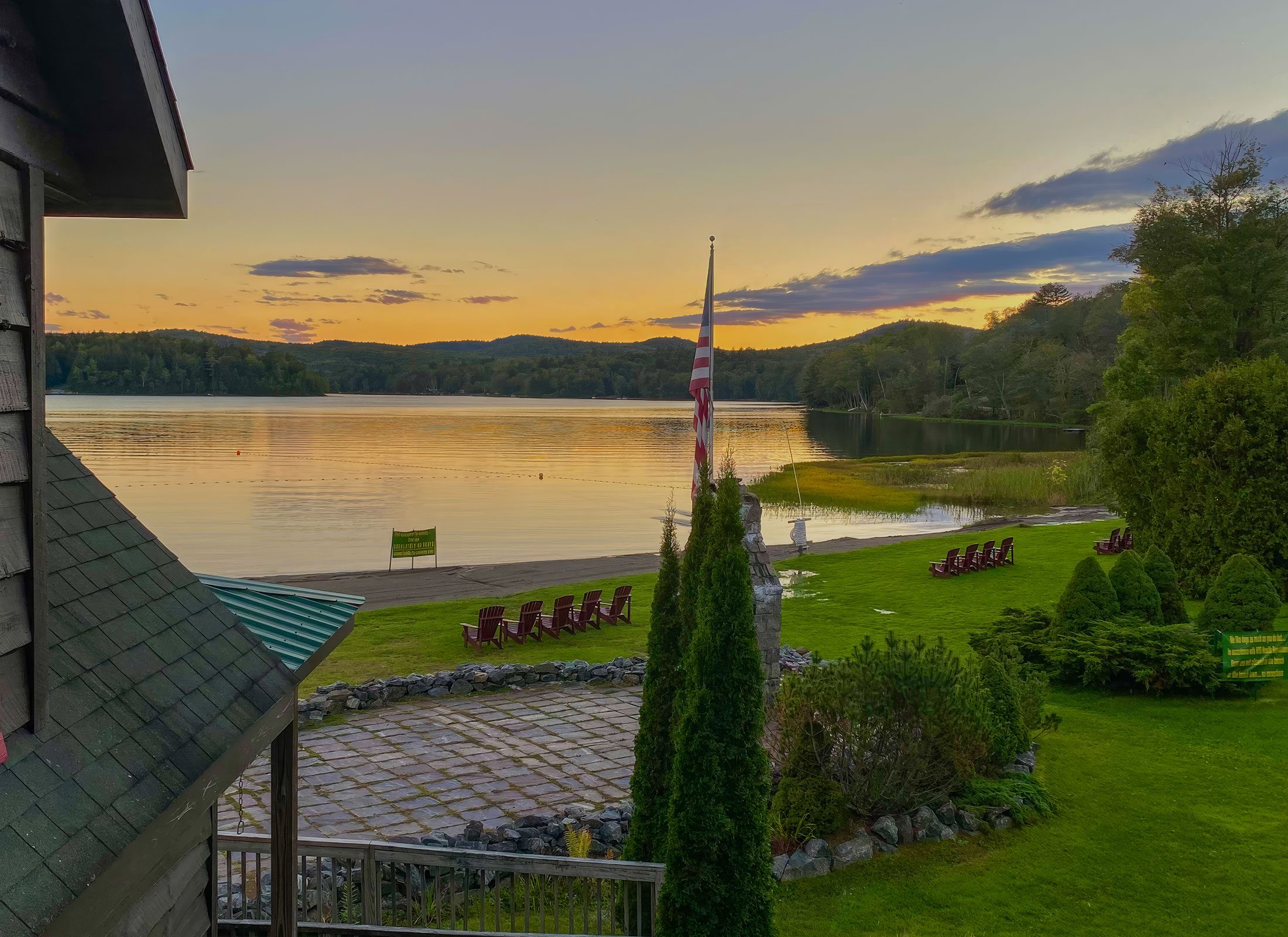 Sunset over Trout Lake from Twin Pines, with the flag, red Adirondack chairs on the lawn and the sandy beach below wooded hills